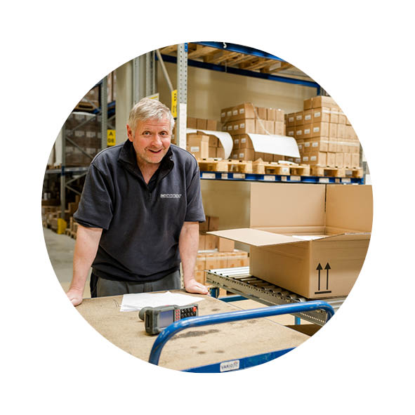 Ein Mann steht im Lager an einem Paketband, er lehnt sich auf einem Wagen ab