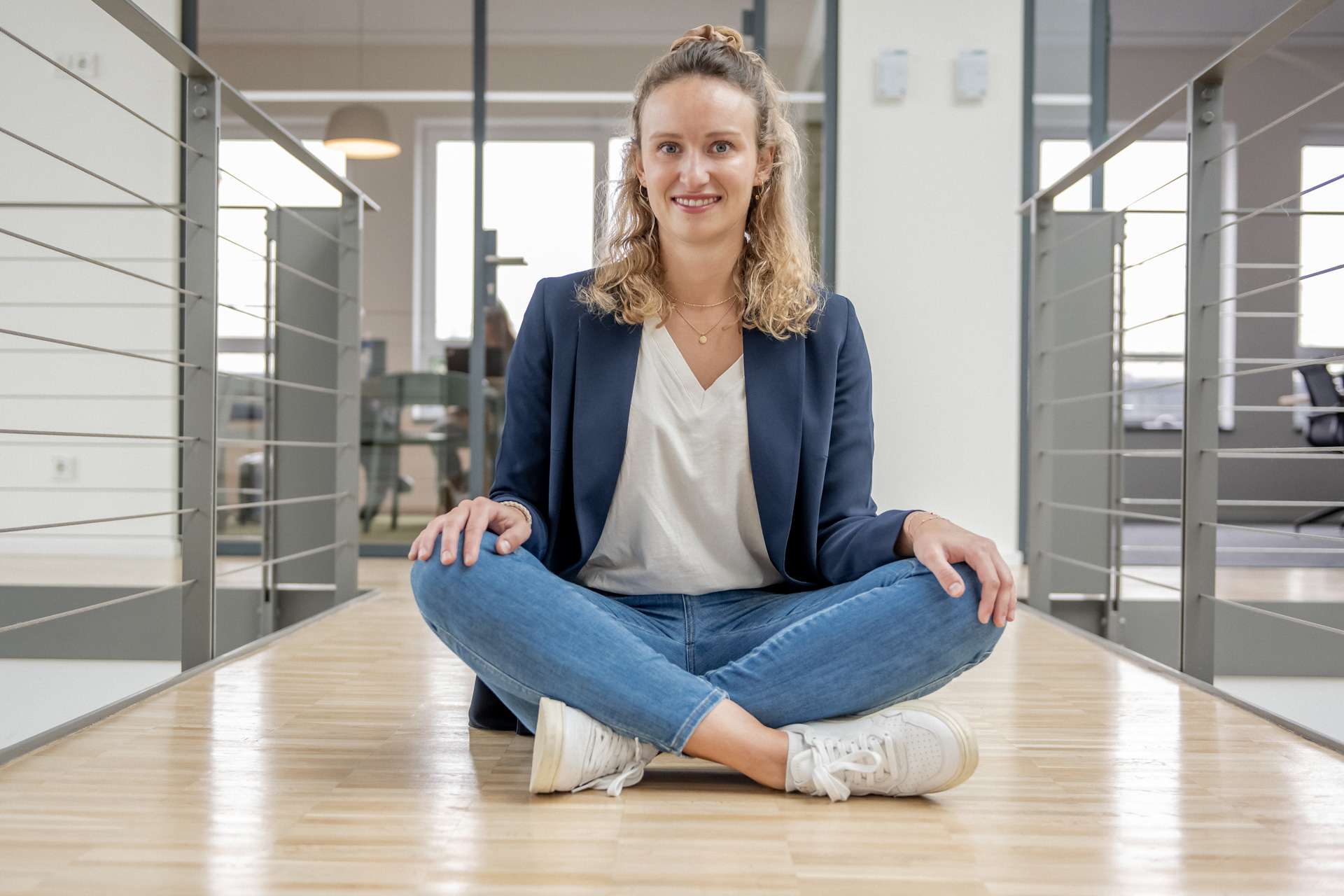 ann-katrin-weidling-weicon Eine junge Frau sitzt im Schneidersitz auf dem Boden in einem Büroflur und lacht in die Kamera
