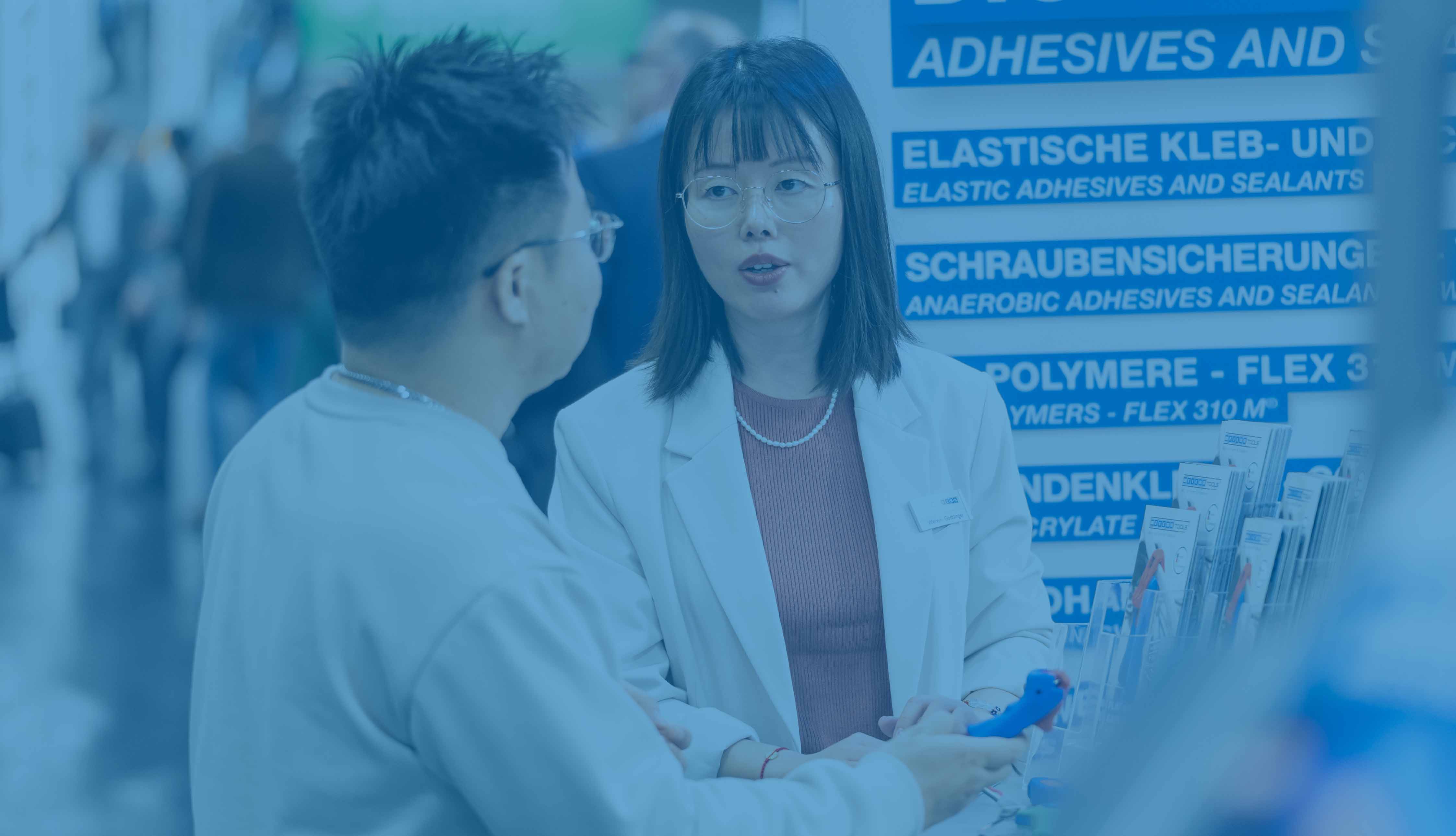 Ein Mann und eine Frau unterhalten sich an einem Messestand, den Mann sieht man von hinten, die Frau trägt eine Brille. Das Bild ist blau hinterlegt