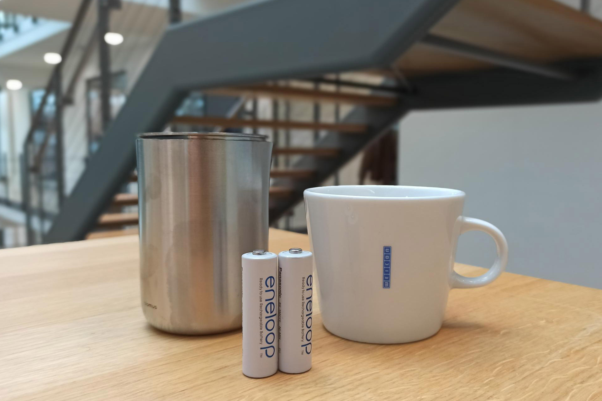 nachhaltigkeit-weicon-massnahmen Ein kleiner Metallmülleimer, Batterien und eine Kaffeetasse stehen auf einem hellen Holztisch