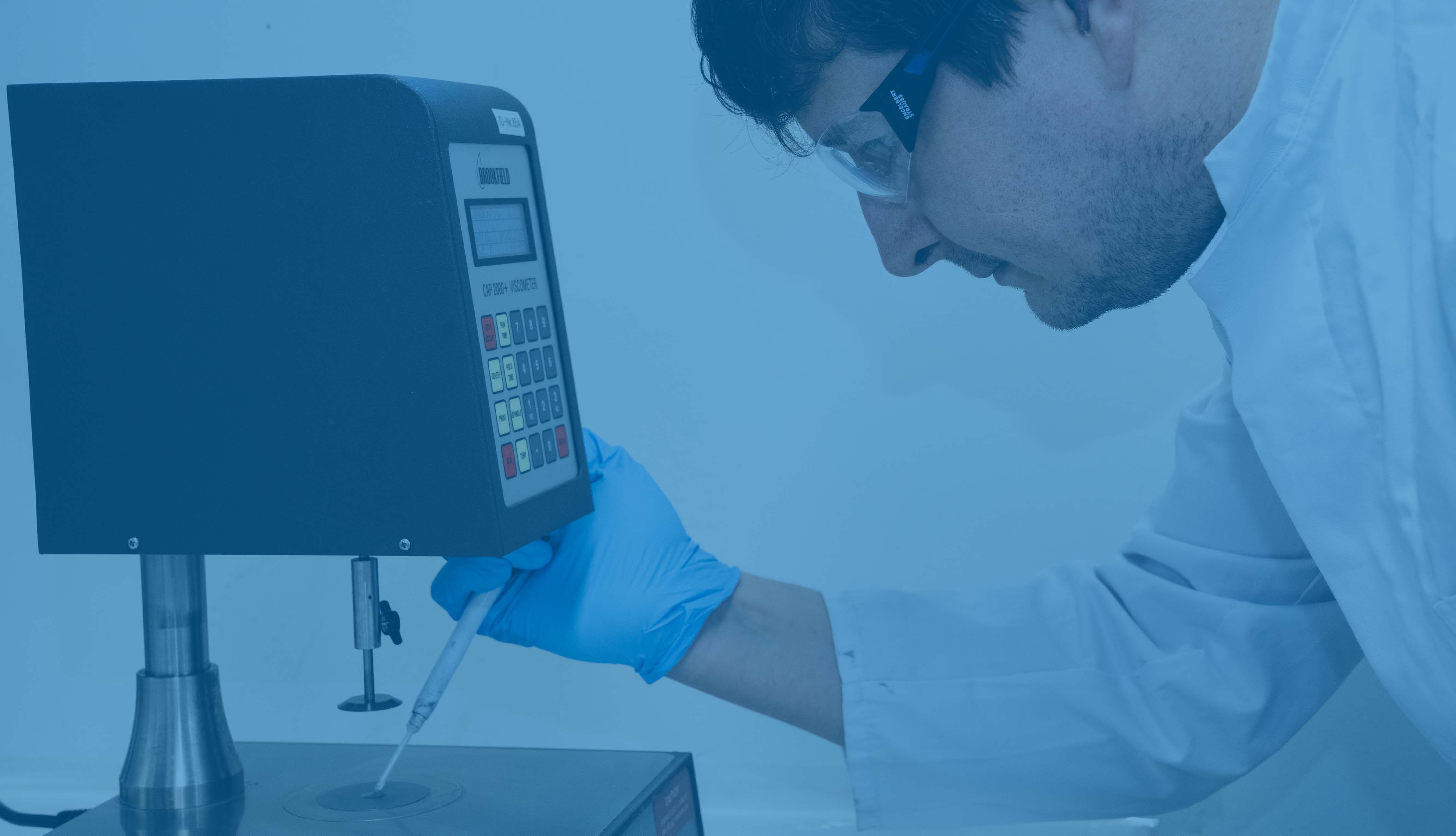 Ein Mann trägt eine Schutzbrille, blaue Handschuhe und einen weißes Kittel und gibt mit einer Pipette etwas auf eine Waage, Bild ist blau hinterlegt