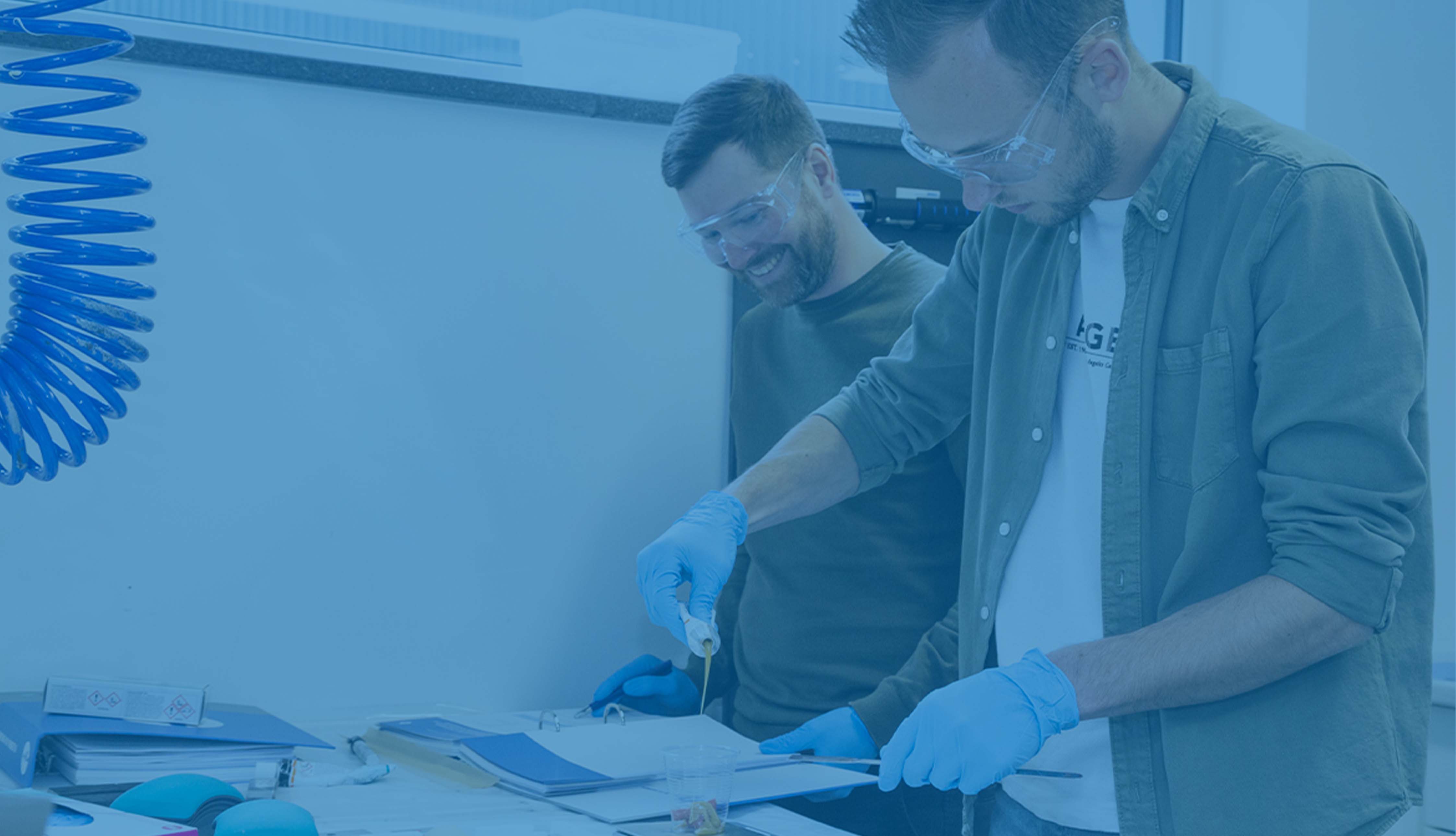 Zwei Männer arbeiten mit Schutzbrille und blauen Handschuhen in einem Labor, Bild ist blau hinterlegt