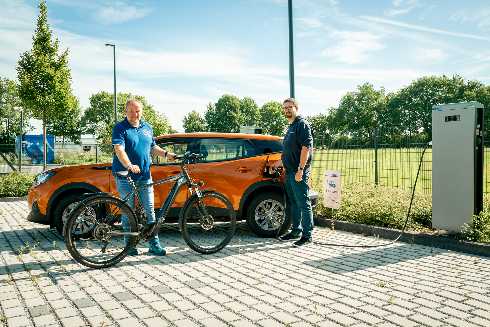 e-mobilitaet-fahrrad-leasing-weicon Ein Mann steht an seinem orangenen Auto das an einer E-Ladesäule angesteckt ist auf einem Parkplatz, ein anderer steht mit seinem Fahrrad davor