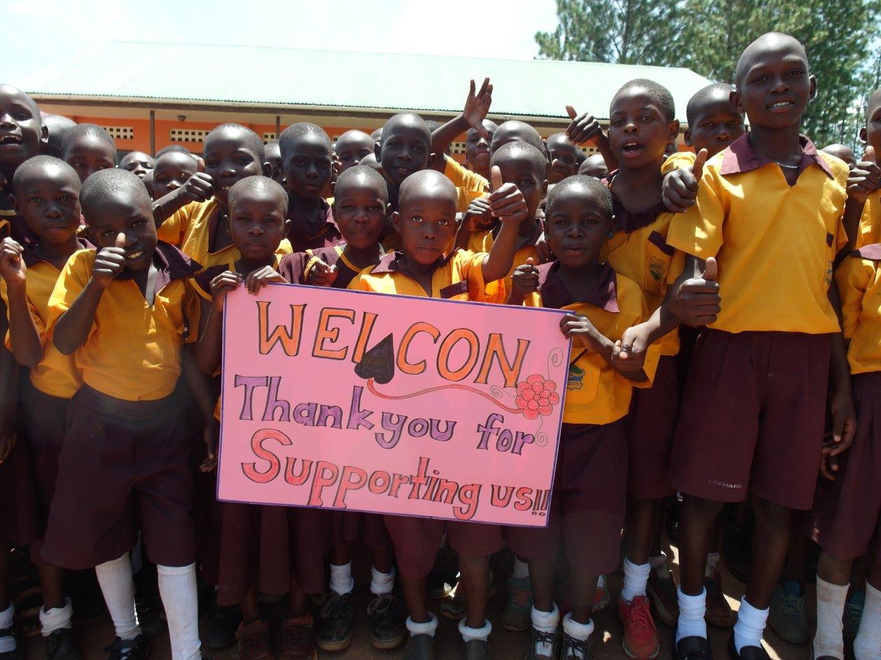 lichtstrahl-uganda-sponsor-weicon eine Gruppe dunkelhäutiger Kinder mit gelben T-Shirts stehen nebeneinander und halten den Daumen hoch. Einige halten ein pinkes Plakat in der Hand auf dem steht: Weicon, thank you for supporting us