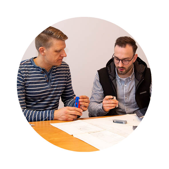 Zwei Männer sitzen an einem Schreibtisch und schauen sich einen Plan an, einer hat ein Werkzeug in der Hand, der andere ein Tablet