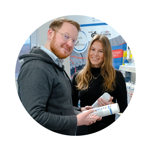 Ein Mann hält eine Spraydose in der Hand und lacht und die Kamera, eine Frau steht daneben hat eine Spraydose in der Hand und lächelt auch in die Kamera, daneben ist ein Regal mit Weicon Produkten